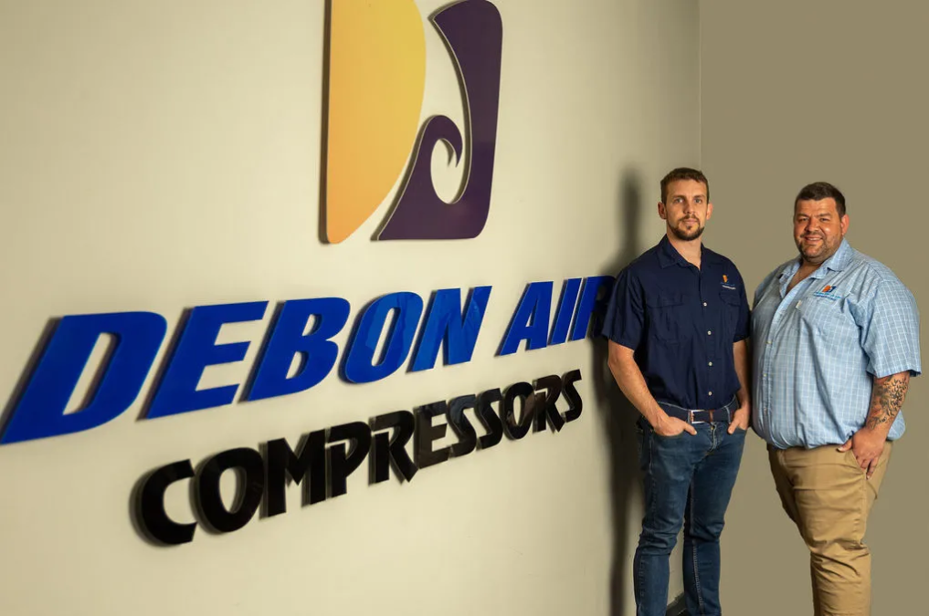 Danie and Jaco Danie standing in front of the Debon Air Compressors logo on the wall at their offices.